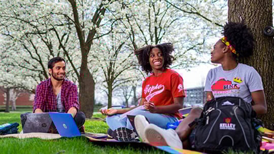 Indiana University students studying outdoors under blooming trees on campus, with laptops and backpacks, representing a diverse and engaged learning environment.
